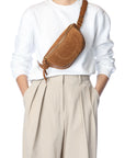 A person in a white sweater and beige pleated trousers carries the handmade Jerome Dreyfuss Lino Banana bag, a brown leather crossbody, across their chest. The photo is set against a plain white background.