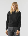 A woman with straight blonde hair wears the Alex Mill Alice Polo Cashmere sweater in black and dark gray jeans, standing against a plain light background with a neutral expression.
