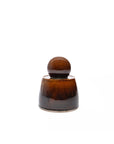 The Faire Amber Stash Jar is a small, brown ceramic container with a glossy amber glaze and a matching rounded lid, shown on a plain white background.