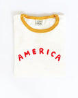 The America Puff Ringer Graphic T-Shirt by Oat Collective is a folded white tee with yellow trim, made from premium Airlume cotton and featuring "AMERICA" in bold red puff print on the front.