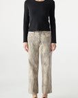 A woman faces forward against a plain white background, wearing a fitted black long-sleeve top, AMO Denim Billie Pants—premium rigid denim cropped wide leg jeans in beige snake-print—and black loafers.