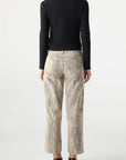A person with long dark hair, wearing a black long-sleeve top and AMO Denim Billie Pants in relaxed-fit beige snake-print, stands facing away from the camera against a plain white background.