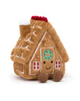 The Amuseables Gingerbread House by Jelly Cat Inc. is a soft, plush toy with white icing details, red windows, a smiling face, and brown feet. Its roof features a chimney topped with white icing and red accents.
