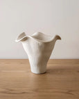 The Faire Andrea Bouquet Vase—a white, textured ceramic piece with an organic, modern shape—rests on a light wood surface against a plain, light background.