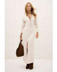 A woman stands against a plain background wearing the BA&SH Dress Anisa—a long, cream ribbed wool blend button-down dress with long sleeves. She pairs it with brown ankle boots, a large brown bag, and has loose wavy blonde hair and minimal jewelry.