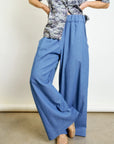 A person wears Aquarius Cocktail’s Connor Wide Leg Pant in blue with a short-sleeved gray camo top and beige studded shoes, standing on a concrete floor. Their face is not visible.