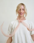 A woman with blonde hair wears an Aquarius Cocktail Dear Polly Bow Blouse against a plain background. A sheer pink scarf is tied around her neck, its ends flowing like mesh tulle. She appears relaxed and smiles gently at the camera.
