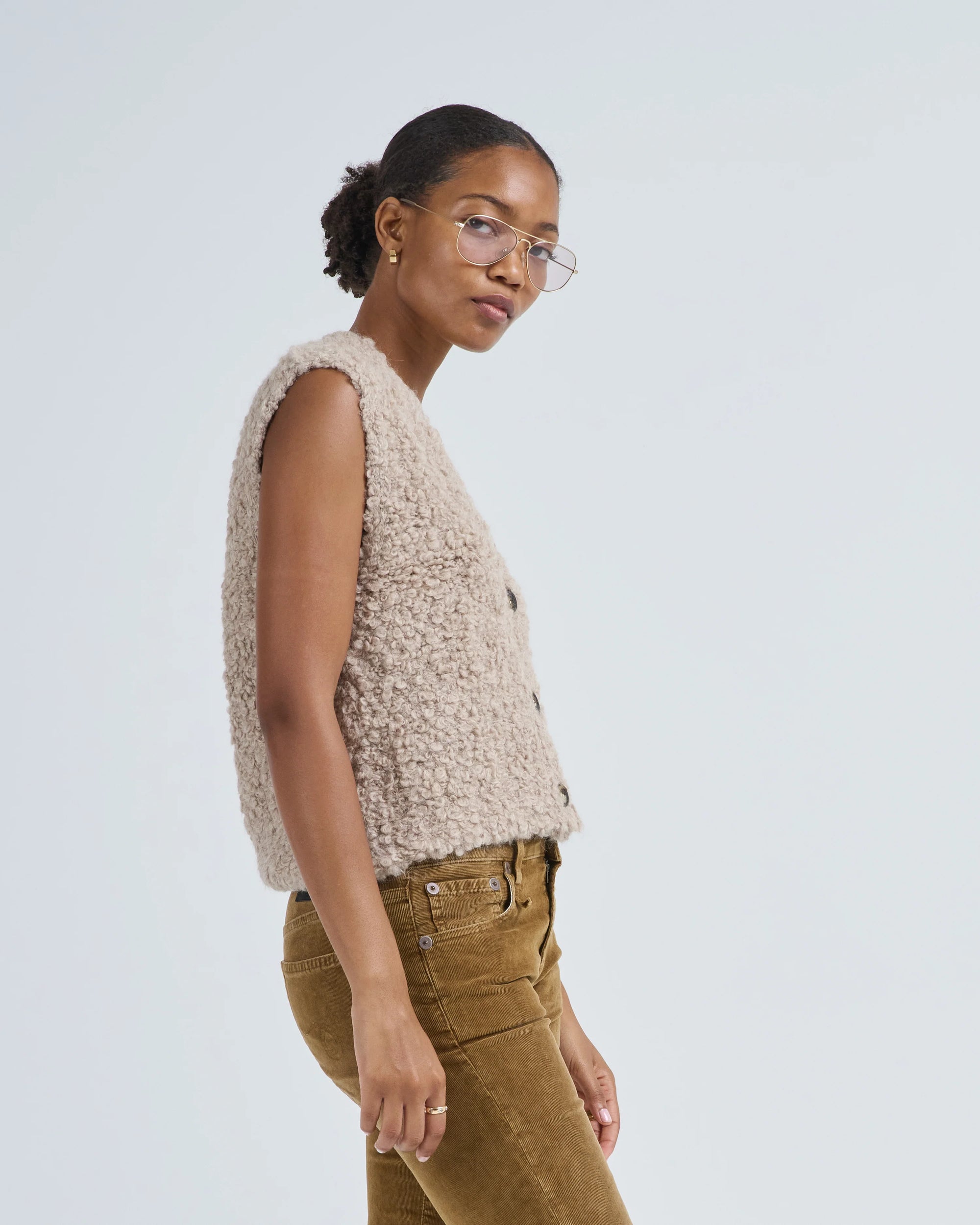 A woman in glasses wears the LaTierra Aries Boucle Vest—an alpaca blend, beige textured sleeveless vest with a V neckline—and brown pants, standing sideways against a plain light background, looking toward the camera.
