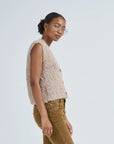 A woman in glasses wears the LaTierra Aries Boucle Vest—an alpaca blend, beige textured sleeveless vest with a V neckline—and brown pants, standing sideways against a plain light background, looking toward the camera.