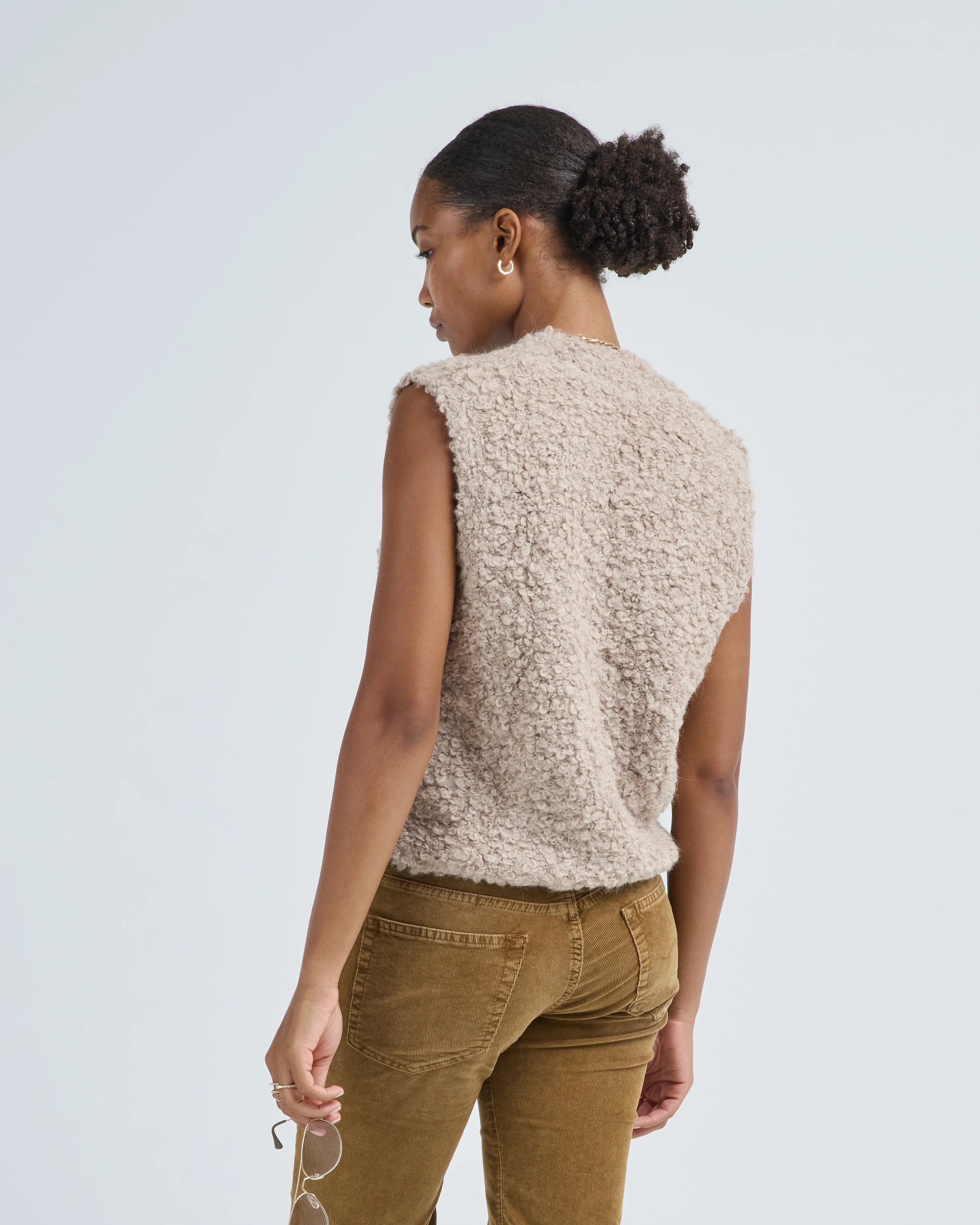 A woman with dark hair in a low puff stands against a light background, wearing the LaTierra Aries Boucle Vest—a sleeveless, textured beige alpaca blend top—with brown corduroy pants and holding eyeglasses in her hand.
