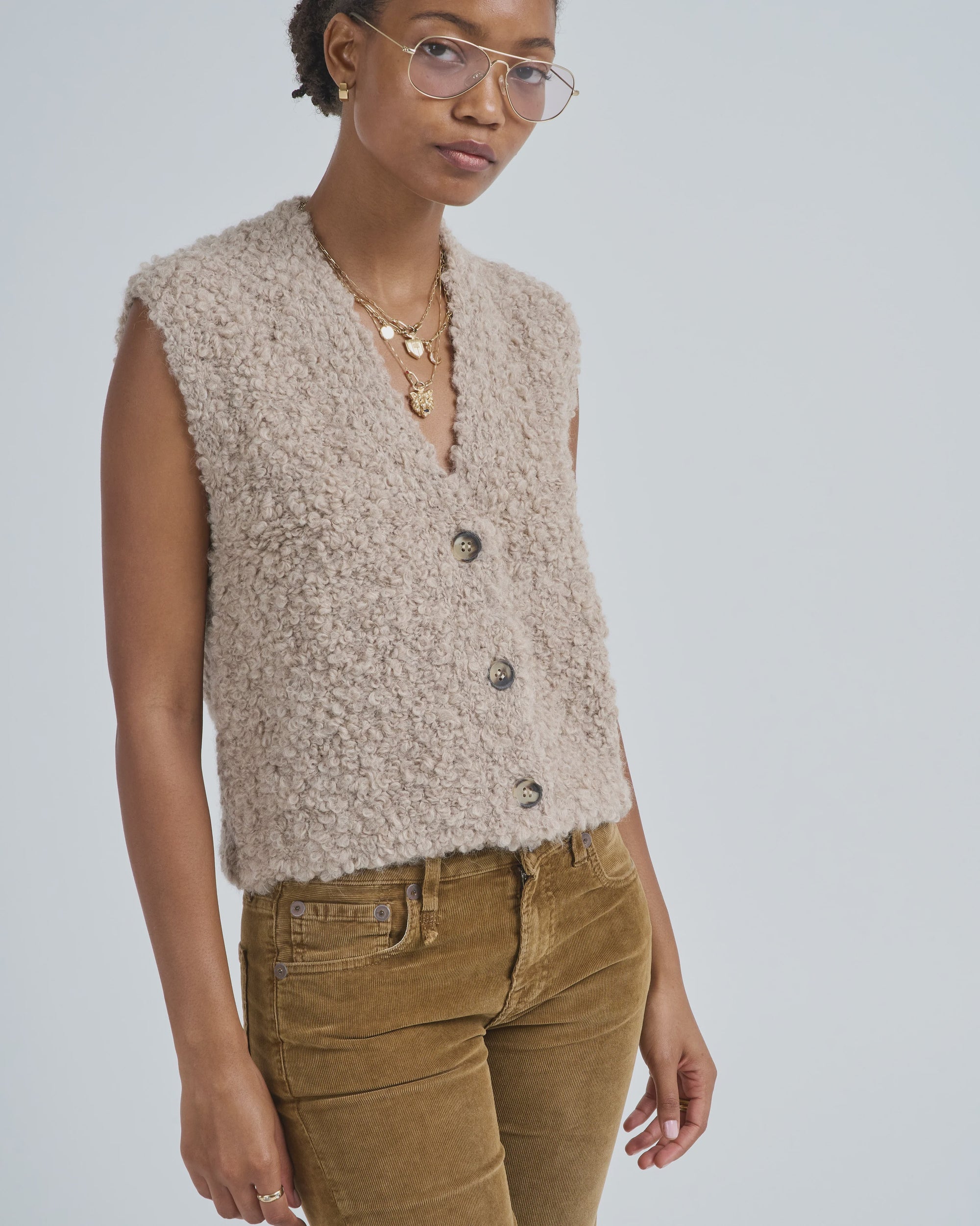 A woman wearing glasses, layered necklaces, brown corduroy pants, and the LaTierra Aries Boucle Vest—a sleeveless beige textured button-up with cow horn buttons and a v neckline—stands against a plain light background, looking slightly to the side.