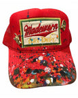 The MadeWorn Hat by Made Worn is a red trucker hat with a mesh back, colorful paint splatters, a custom "Madeworn ART DEPT." front patch, and limited edition pins including a gold hand and small circular pin on the front.