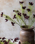 Abigail Ahern’s Cosmos/Burgandy features long green artificial stems with dark burgundy blooms in a rustic brown ceramic vase. Hand-painted fabric flowers are arranged on a light surface, set against a textured beige wall.