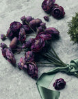 The Abigail Ahern Ranunculus Bunch Purple, featuring vibrant purple blooms with green stems and a green satin ribbon, rests on a gray stone surface, with extra artificial flowers and greenery scattered nearby.