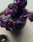 A close-up of the Abigail Ahern Ranunculus Bunch Purple artificial flowers arranged in a vase and viewed from above, with a blurred cup or bowl nearby on a light, round surface.