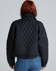 A person with red hair, seen from behind, wears the ASKK Bleecker Street Liner— a black, quilted, cropped jacket—paired with blue jeans against a plain light gray background.