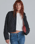 A person with long, wavy red hair wears the ASKK Bleecker Street Liner—a black quilted, cropped jacket with one side open to show its pink lining—paired with a white crop top and blue jeans against a plain light gray background.