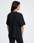 A person with straight brown hair is seen from behind, wearing the ASKK Boy Tee 26, a loose black t-shirt made in Los Angeles, paired with dark jeans against a plain white background.