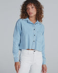 A woman with curly hair models the ASKK Cropped Denim Shirt in light blue with a vintage feel, paired with high-waisted white pants, standing against a plain light gray background.
