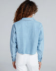 A person with curly hair is seen from the back in an ASKK Cropped Denim Shirt paired with high-waisted white pants, creating a subtle vintage vibe against a plain light gray background.