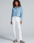 A woman with curly hair wears the ASKK Cropped Denim Shirt in light blue, paired with white high-waisted wide-leg pants and gray sandals. She stands against a plain light gray background, gazing off to the side for a vintage vibe.