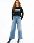 A woman with long curly red hair stands against a white background, wearing an ASKK printed cropped long sleeve shirt, high-waisted wide-leg blue jeans, and black heeled ankle boots.