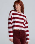 A red-haired person wears the ASKK Cropped LS Tee—long-sleeved, wide maroon and white stripes, soft cotton—paired with high-waisted maroon pants, standing against a plain background.