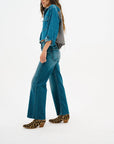 A woman with long brown hair stands in profile against a white background wearing an ASKK Crosby Flannel shirt in pink plaid, a blue denim jacket, blue jeans, and leopard-print low-heel ankle boots.