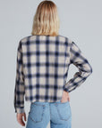 A person with shoulder-length blonde hair, seen from the back, wears the ASKK Crosby Flannel in blue and beige plaid with light blue jeans. Their left hand rests on their hip against a plain light gray background.