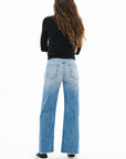 A person with long wavy hair, wearing a black long-sleeve top, ASKK Mercer Jean high-rise wide-leg denim, and black sneakers stands facing away from the camera against a white background.