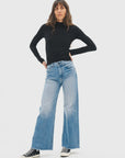 A woman poses against a plain background wearing a black long-sleeve turtleneck, ASKK Mercer Jean high-rise light blue wide-leg Japanese denim jeans, and black-and-white sneakers, her hands resting on her hips.