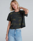 A woman with short blonde hair wears the ASKK Shrunken Boy Tee in dark camo print with light blue jeans, standing against a neutral background and resting one hand behind her neck.