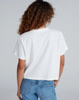 A person with curly brown hair is shown from the back, wearing an ASKK Shrunken Boy Tee and blue jeans against a light gray background.