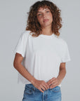 A person with curly brown hair wears the ASKK Shrunken Boy Tee and blue jeans, standing against a light gray background. They face the camera with a slight smile, hands loosely clasped.