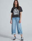 A woman faces the camera in an ASKK Shrunken Boy Tee—a black "New York" crewneck—paired with light blue wide-leg jeans with rolled cuffs and black sneakers with white soles, set against a plain light background.