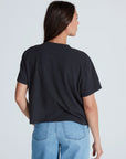 A woman with long brown hair wears the ASKK Shrunken Boy Tee and light blue jeans, standing with her back to the camera against a plain, light background.