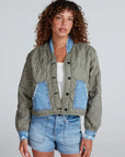 A woman with curly hair wears ASKK’s The Peak Liner, a green lightweight quilted jacket with blue denim collar, cuffs, and pockets, over a white top and denim shorts. She stands against a plain light background.