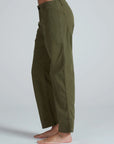 A person stands barefoot against a plain light gray background, wearing ASKK's high-waisted Tomboy Chino in olive green with visible side and back pockets. The loose-fitting, midweight pants are photographed from the side.