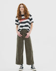 A person with long curly red hair wears a black-and-white striped tee, ASKK’s Virginia Corduroy olive green cropped wide-leg pants, and black high-top sneakers, standing confidently against a white background.