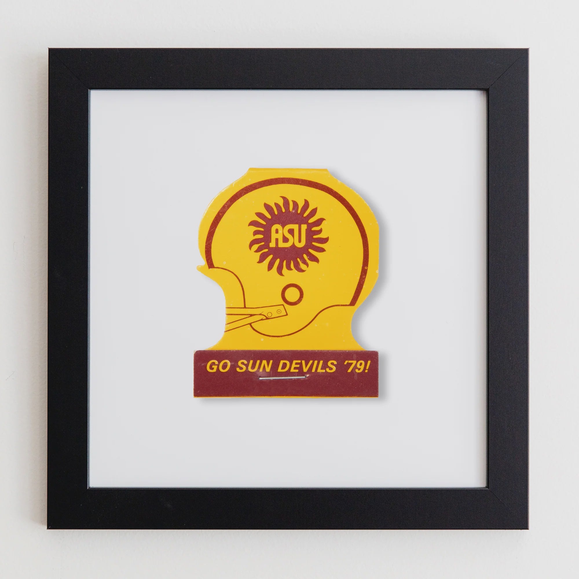 A framed acrylic image featuring a vibrant yellow and orange sunburst design inside a silhouette of a helmet, with the text "GO SUN DEVILS 79!" on a beige background in the Art Square Black Frame by Match South.
