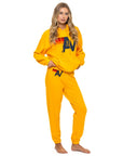 A woman with long blonde hair models the Aviator Nation women's sweatpants, featuring a bold "AV" logo and red accent, in bright yellow. She stands barefoot against a plain white background, channeling a vintage vibe.