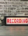 Faire's Lighted Sign features bold red "RECORDING" letters illuminated on a classic marquee, resting on wood near a vintage fan and brick wall—ideal personalized wall decor for creative spaces.