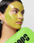 A woman with wet, dark hair wears a shiny green Loops/HydroGel Face Mask by Faire and silver hoop earrings. She holds bright green packaging with black text near her face, looking left against a plain background.