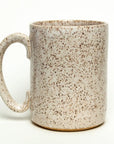 The Faire Ceramic Mug features a large handle and speckled beige and brown glaze. Handmade, dishwasher safe, and ideal for everyday use, it’s shown against a plain white background.