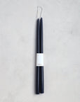 The Floral Society’s 18" Taper Candle - Pair features two tall, slender dark blue candles joined by a wick and wrapped with a white “CANDLE” label, ideal for elegant home décor. Displayed on a light gray textured surface.