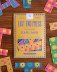 A yellow and blue Faire Paper Garland/Chains box rests on a wooden surface, surrounded by vibrant birthday garlands and paper chains in pink, blue, and yellow with floral and geometric designs.