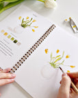 A person uses a paintbrush to paint yellow tulips in the Faire Spring Watercolor Workbook, surrounded by paint tubes, a palette, and a fresh tulip—ideal for beginner art activities on a white surface.