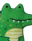 The Faire Alligator Embroidery Kit features a green fabric crocodile plush with embroidered eyes, teeth, and colorful patterns inspired by alligator designs, displayed against a white background.