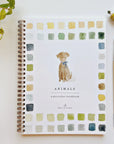Faire's "Watercolor Workbook," titled "Animals," rests on a surface. It features beginner-friendly illustrations and a spiral binding for easy page turns. The cover, with vibrant squares, displays a painted dog with a blue bow. Nearby lie a paintbrush, paint tube, and green-leafed plant.