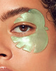 Close-up of a person with curly hair, a green tropical leaf-shaped gel patch from Faire's Cucumber/Tea Eye Mask is applied under their right eye. Their skin looks dewy and moisturized.